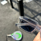 A close-up of a person holding a pair of translucent clear-framed sunglasses with dark lenses, with a blurred outdoor court background and a paddle visible on the ground.