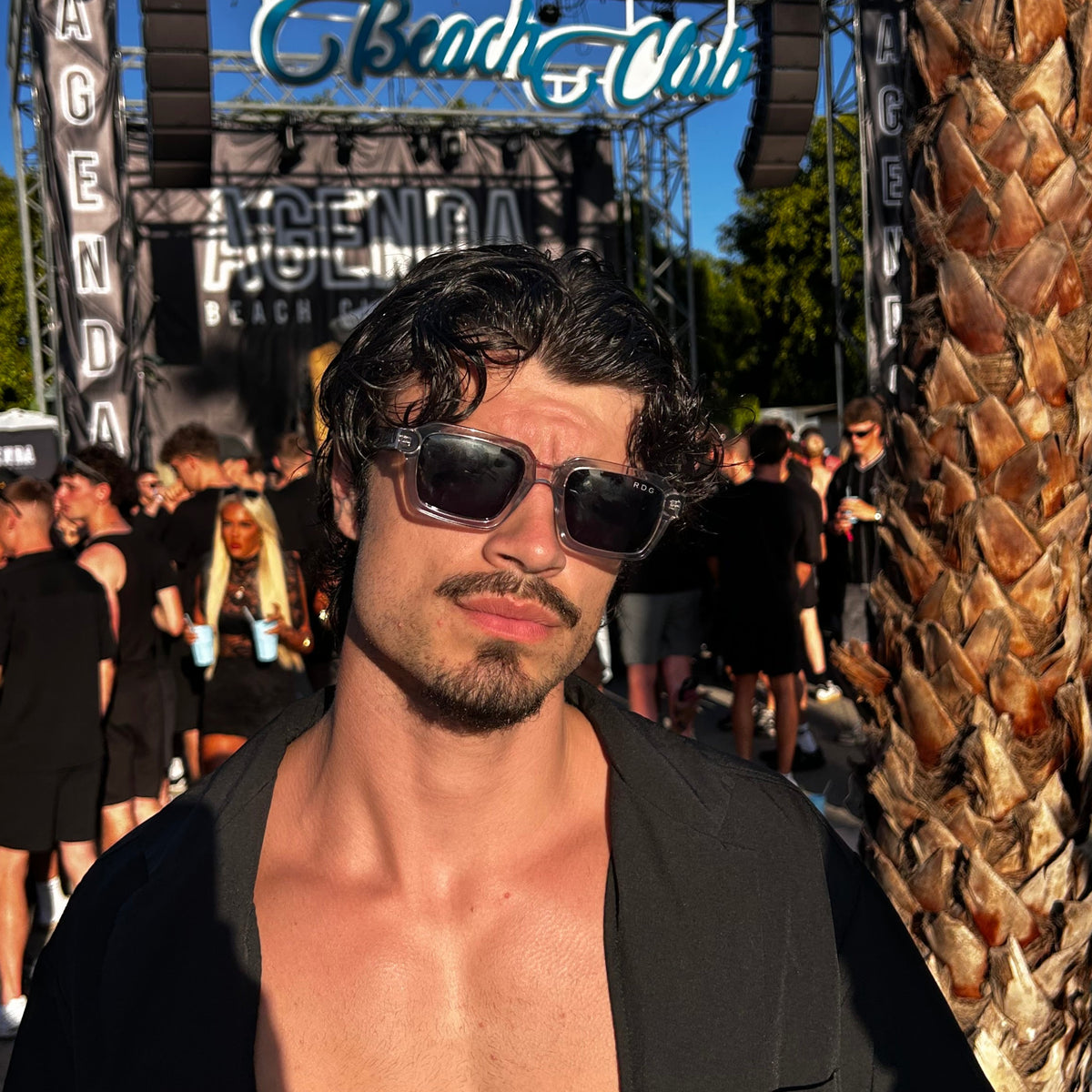 A shirtless man wearing clear sunglasses and an open black blazer stands in front of a poolside event backdrop with a large 'Heaven Beach Club' sign and a palm tree at night.