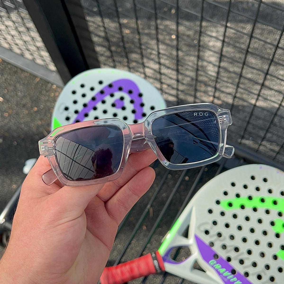 A hand holds a pair of clear-framed sport sunglasses with dark lenses in front of a black wire fence, with a white and purple padel racket and a red grip handle visible in the background.